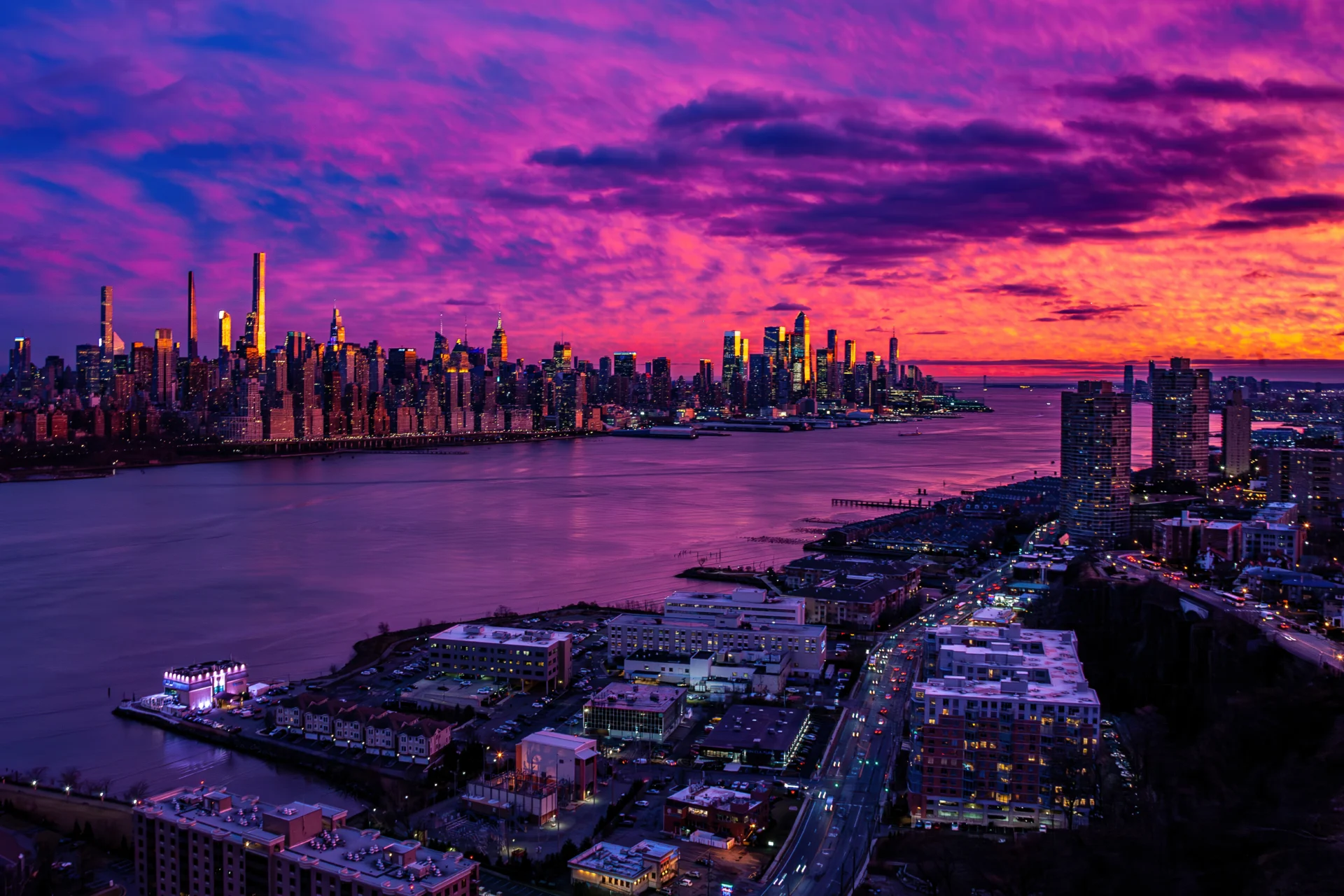 Dramatic purple and orange sunset sky over a vibrant city skyline reflecting in a wide river with waterfront buildings.