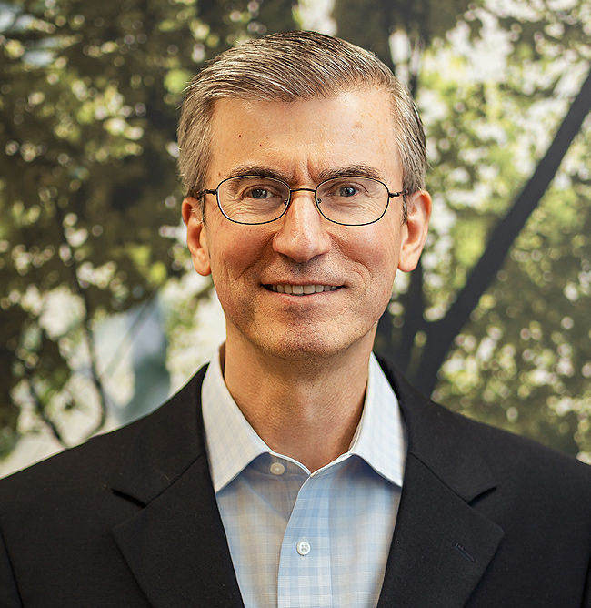 Daniel Vira, a smiling man with gray hair, glasses, blue plaid shirt, and dark jacket against blurred green foliage.