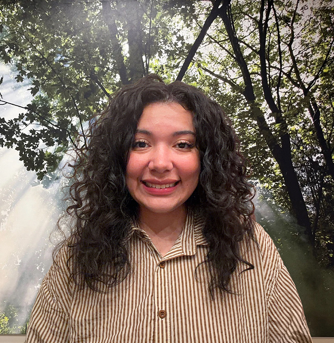 Taylor Lopez, a smiling woman with dark curly hair, wears a striped shirt against a sunlit forest background.